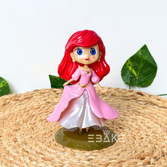 Figurine of a character with red hair and a pink dress on a woven surface with leaves in the background.